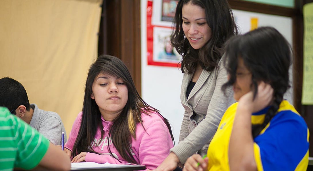 Jasmine Juarez stands over two girls who are sitting next to each other at desks, and Juarez offers input on the math assignment they are working on.
                  