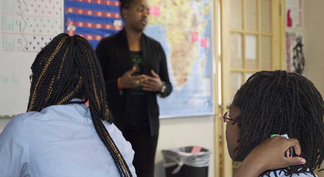 Teacher Dayo Harris stands at the front of a classroom, in front of a whiteboard and a map of the continent of Africa, delivering a lesson to a classroom of students.  In the foreground of the image, two Black female students are listening to Harris talk.
                  