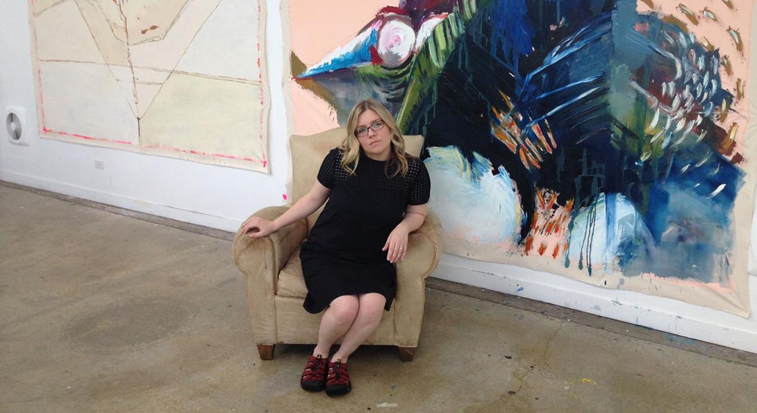 Rachel Harper sits in an easy chair in front of a large work of art, an abstract painting, in an art gallery.