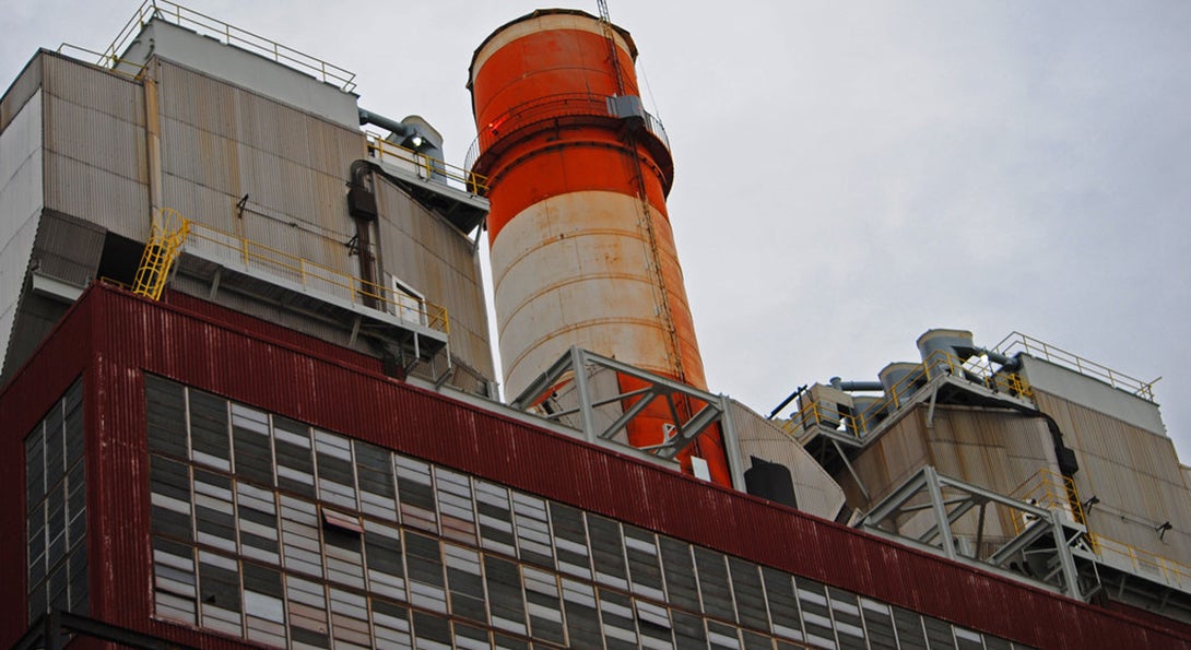 The Crawford Generating Station, a now-retired coal plant in Chicago.  A rusted orange smokestack rises above a series of boarded up window panes on a large red brick building.
                  