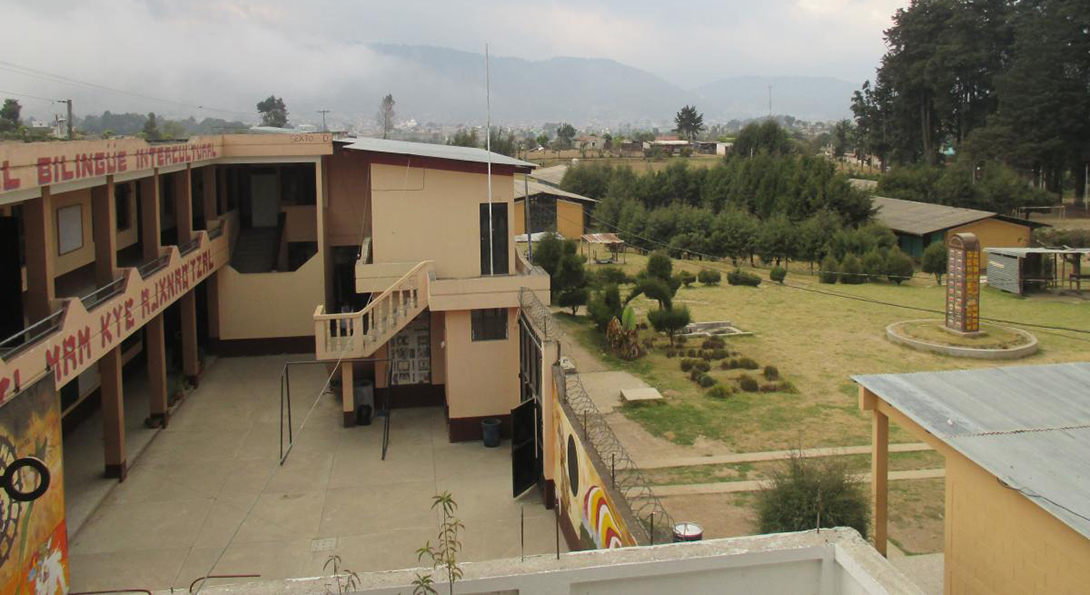 A school building and its courtyard in Guatemala, with mountains in the background and low clouds and fog shrouding the view of the mountains.
                  