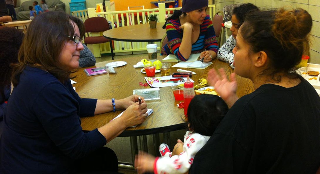 Professor Norma Lopez-Reyna records the story of a woman at the Maria Shelter in Chicago.  The woman telling her story is sitting at a table and holding her baby in her lap.
                  