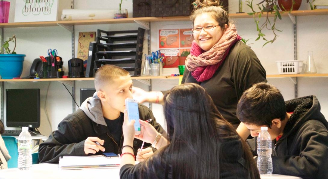 Teacher Adilene Aguilera stands by a group of three students sitting at a table, discussing their current science experiment with them.