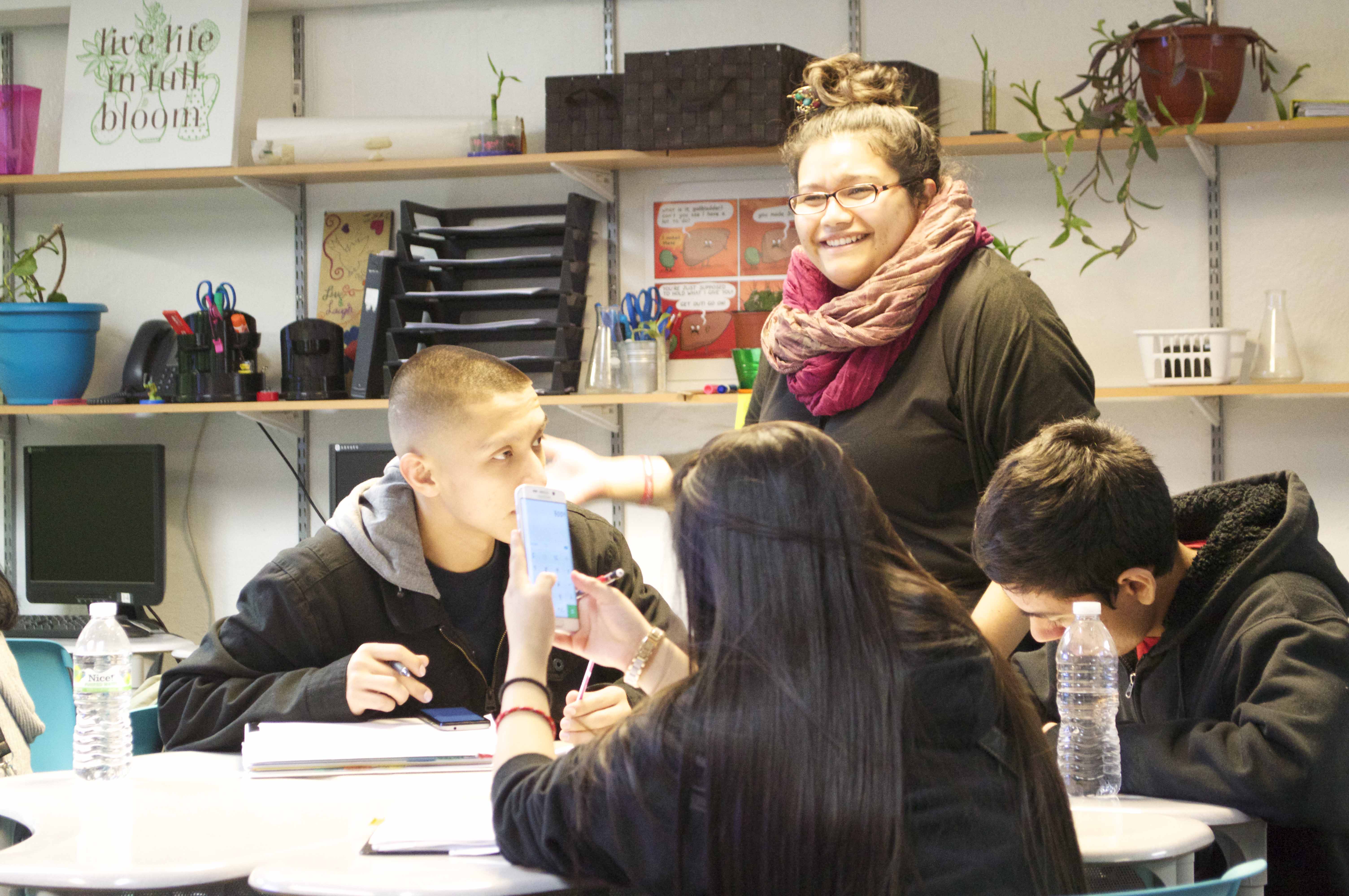 Teacher Adilene Aguilera stands by a group of three students sitting at a table, discussing their current science experiment with them.
                  