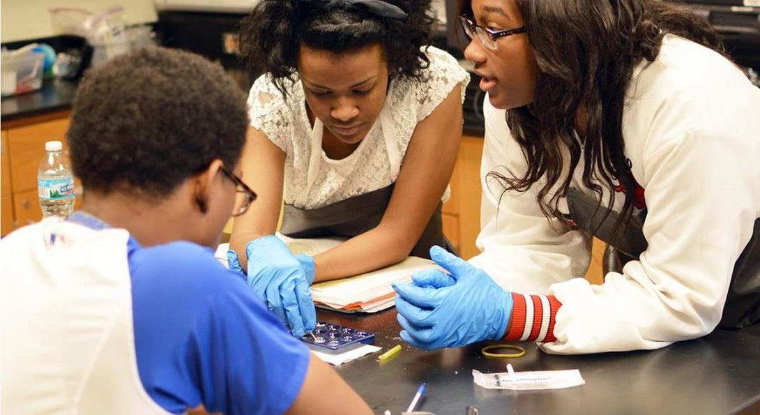 Three Black students are working on a science experiment at a table in a chemistry classroom, moving small amounts of liquid from one container to another.