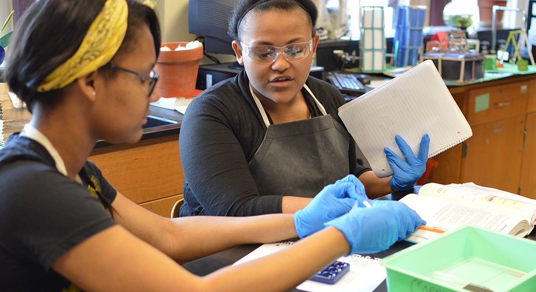 Two Black girls work on a science experiment, removing liquid from a vial and transferring it to a beaker.