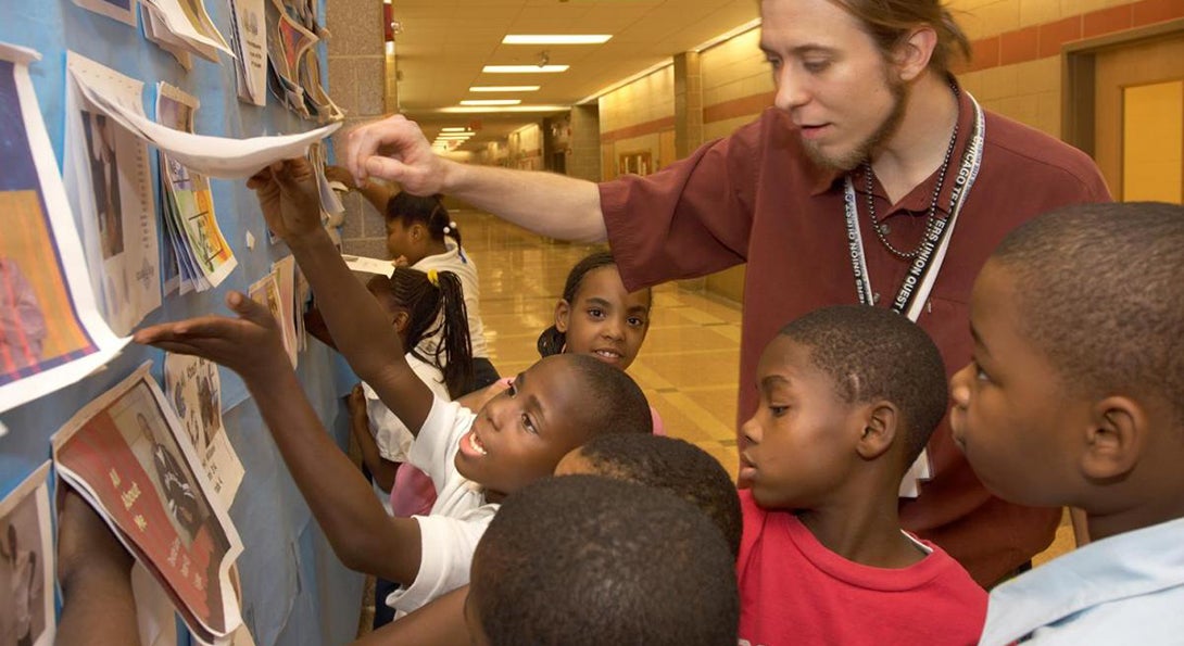 A teacher is looking at examples of student art on a bulletin board in a school hallway, surrounded by young Black male students.
                  