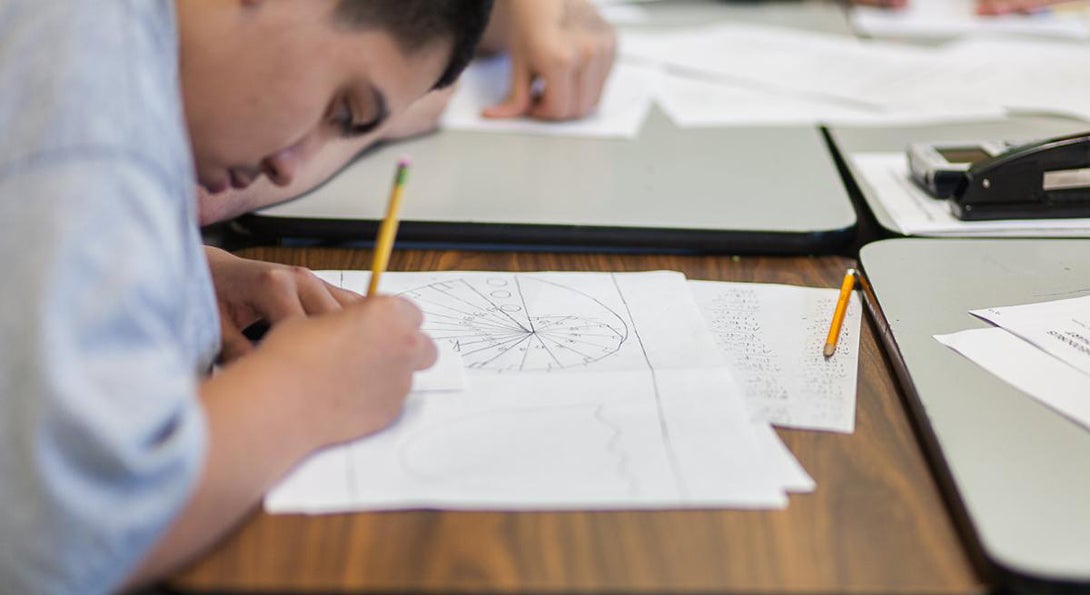 A Latino student works on a math problem with pencil and paper sitting at a desk in a classroom.
                  