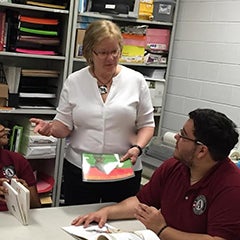 Maureen Meehan stands in a small classroom with three students, who are sitting at a table reading books.  Meehan is having a conversation with one student about the book.