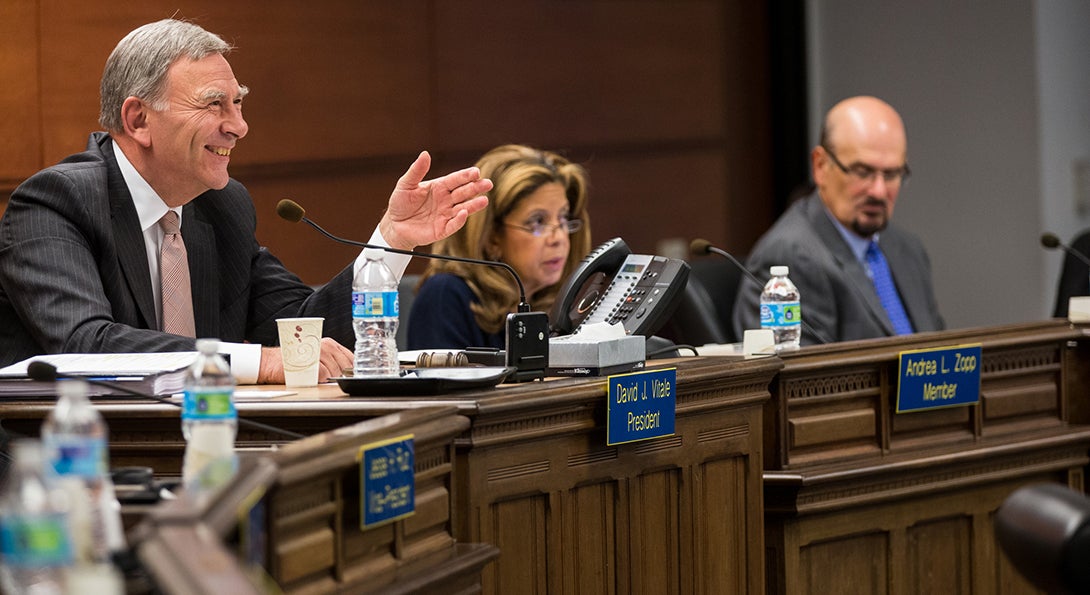 At a Chicago School Board meeting, board president David Vitale gestures to a speaker who is addressing the board, while two other board members are looking on at the interaction.