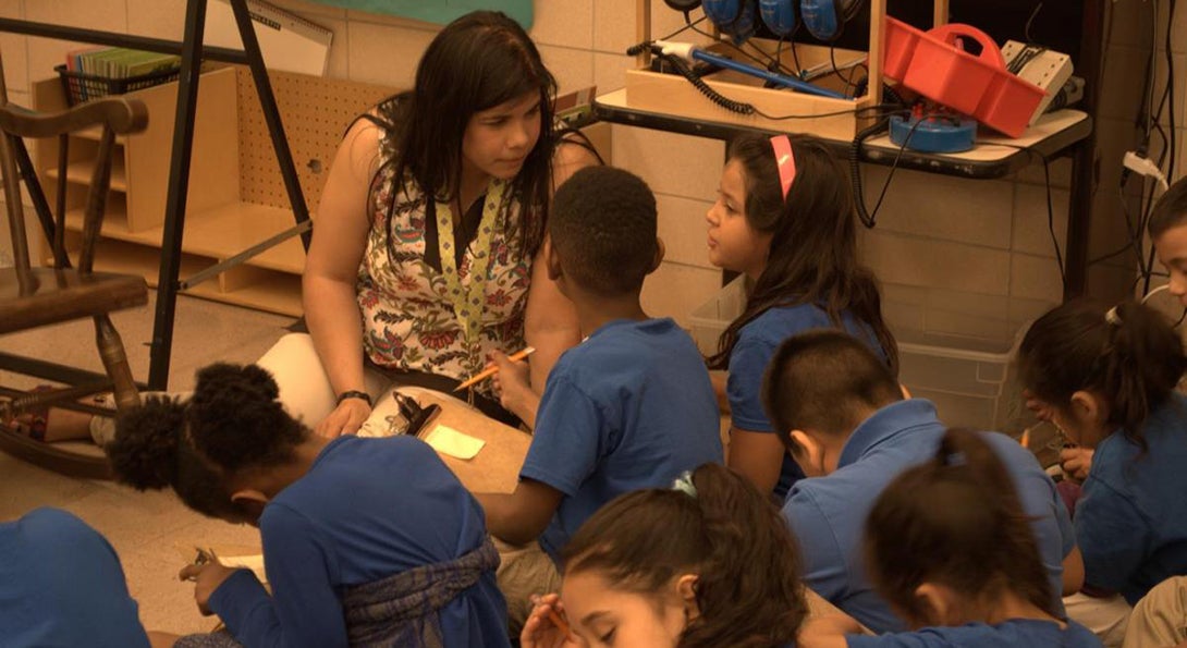 A teacher sits on the floor and talks with two Latino students who are working on an activity together, writing down responses on a clipboard.
                  