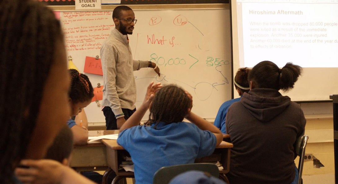 Evan Taylor teaches a math class, pointing to numbers and figures written on a whiteboard.  Students in his classroom are sitting at desks and listening to the lesson.
                  