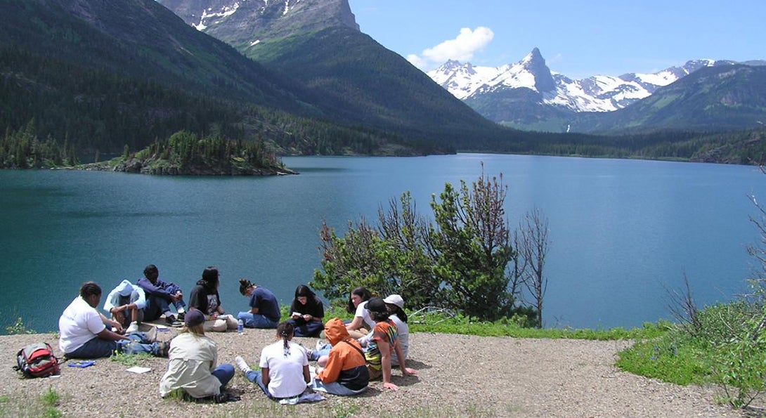 Girls sit in a circle on a sandy beach beside a lake surrounded by mountains as part of a Project Exploration trip.
                  