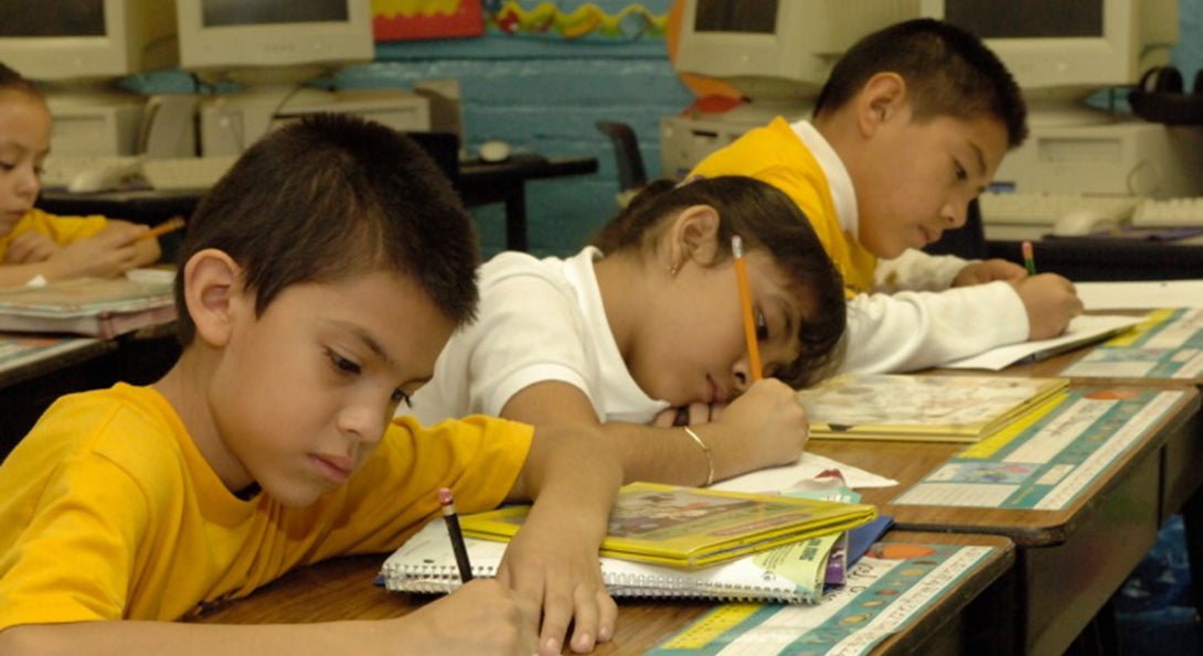 Three Latino students sit in a row at desks, each working on writing in workbooks.