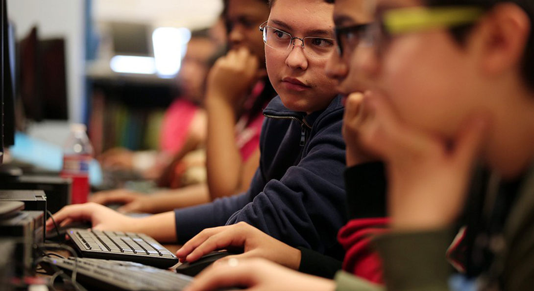 At Wells Community Academy High School, students sit along a row of computers, each working on a coding task during the Hour of Code event. One student is looking at the screen of the student sitting to his left.
                  