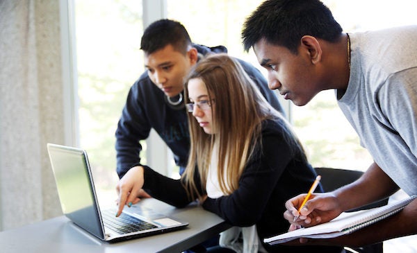 Three students are working on a group project, with a woman sitting working on a laptop and two male students standing beside her on either side, looking at the laptop and taking notes in a notebook.