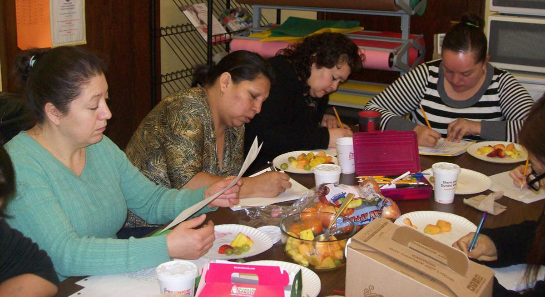 A group of Latina mothers sit around a table, eating lunch and working on writing stories on memories from their lives.  One woman is holding papers in her hands, reading her work.