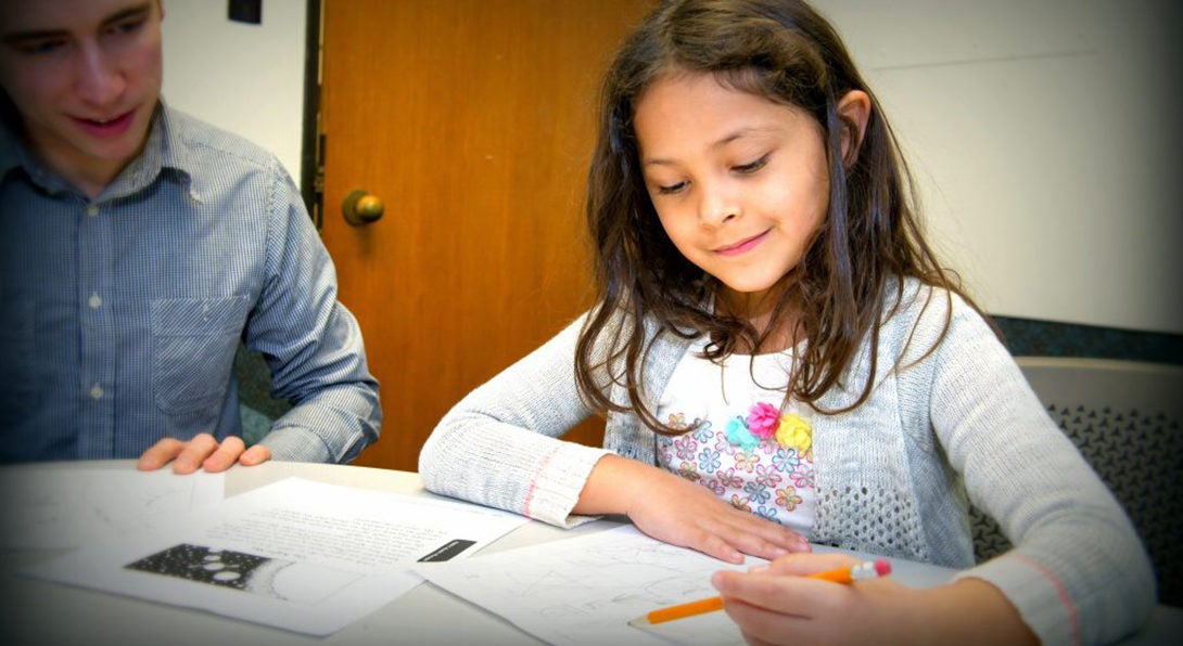 A girl undergoes an education assessment, working through a series of problems on a piece of paper while holding a pencil.  A Masters of Special Education student is sitting to her right observing her work.