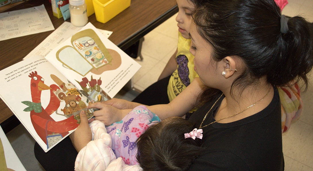 At a Center for Literacy workshop for ESL parents, a Latina woman reads a children's books to her two kids, who are both sitting in her lap.