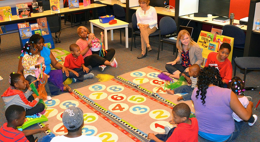 Doctoral student Colleen Whittingham leads a workshop with young mothers focused on developing literacy skills for early childhood learners.  The group is sitting in a circle on a colorful rug featuring numbers, and they are singing a song about counting.