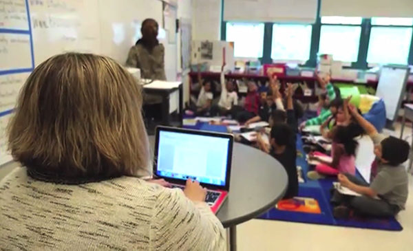 A woman sits at a table with a laptop, entering data from observations she is conducting.  She is observing a teacher in action in a classroom.  The teacher is writing on a whiteboard, while the teacher's students sit on a rug in a circle around her.  Several students have their hands raised.