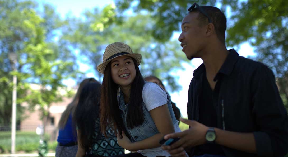 Students sit on a bench under a tree on campus, having a conversation.