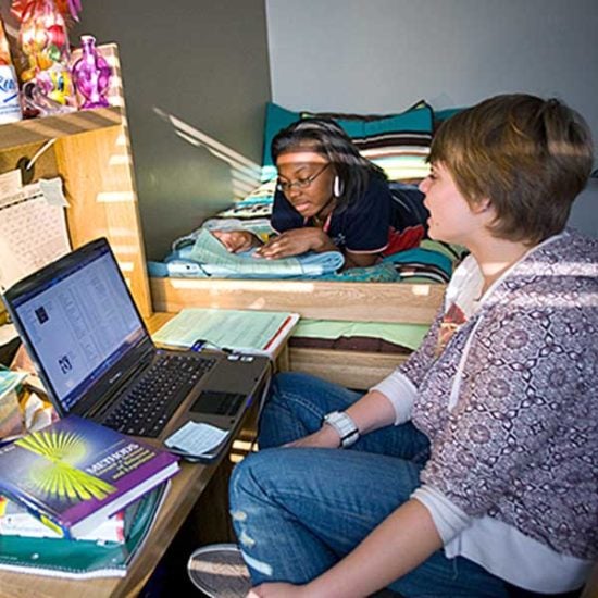 A student sits at her desk in a dorm room, looking at her computer, talking to a girl laying on a bed to her right.