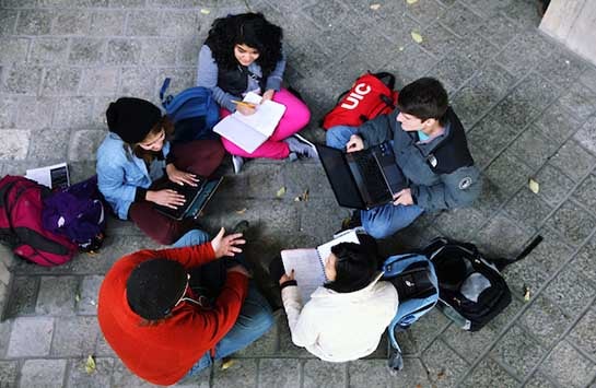 Five UIC students sit in a circle, talking and typing on laptops.  They are on the patio atop the BSB building on UIC's campus.