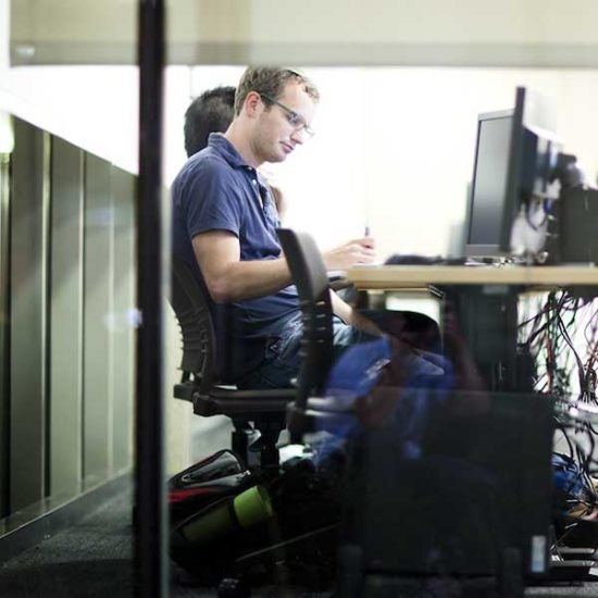 A student sits at a computer in a computer lab on campus, writing on a piece of paper and looking at his computer screen.