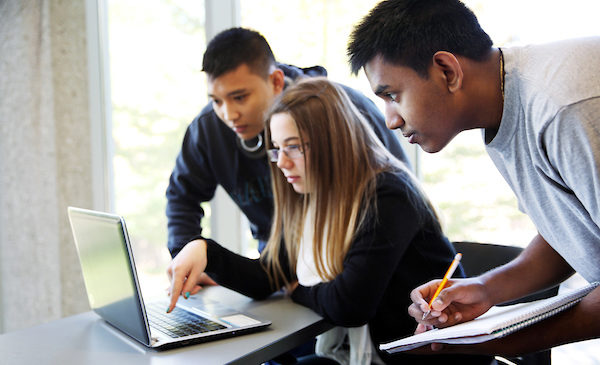 A female student sits looking at a laptop, while two male students are standing behind her, having a discussion and taking notes in their notebooks.