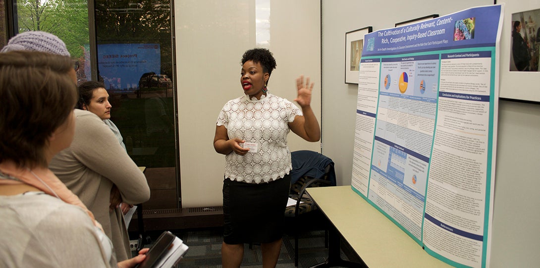 Master Teaching Fellow Mindy Chappell stands in front of her poster, explaining her research to a group of Project SEEEC fellows standing in front of her.