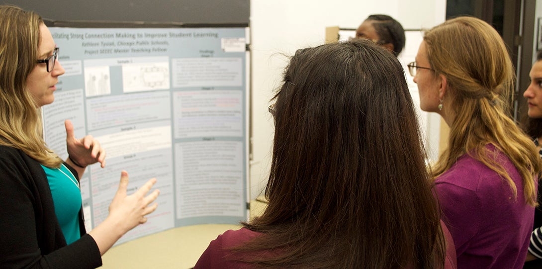 Master Teaching Fellow Kathleen Tysiak stands in front of her research poster, explaining her findings to peer fellows standing in front of her.