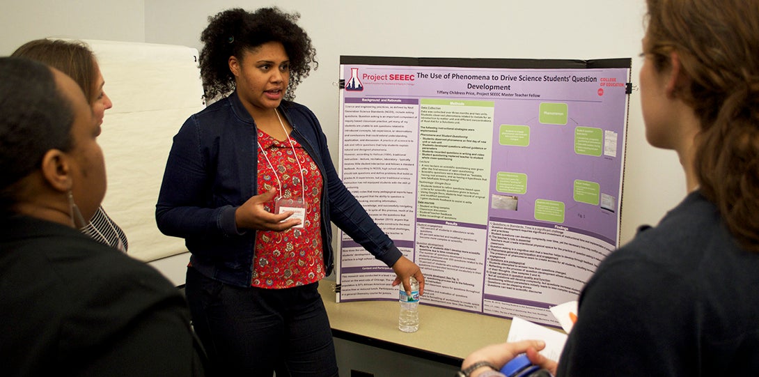 Master Teaching Fellow Tiffany Childress Price stands in front of her research poster, explaining her findings to peer fellows standing in front of her.