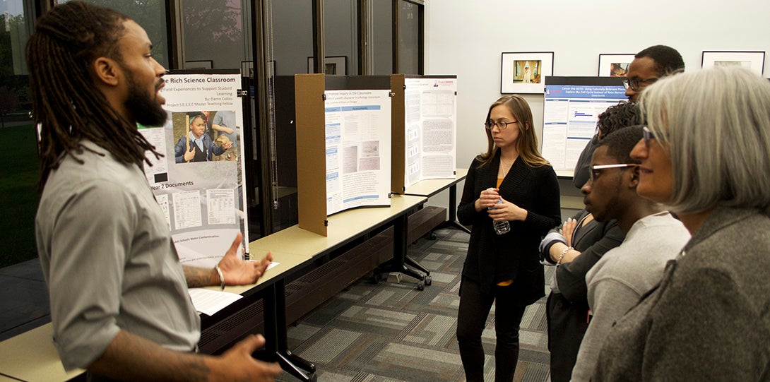 Master Teaching Fellow Darrin Collins stands in front of his research poster, explaining his findings to peer fellows standing in front of him.