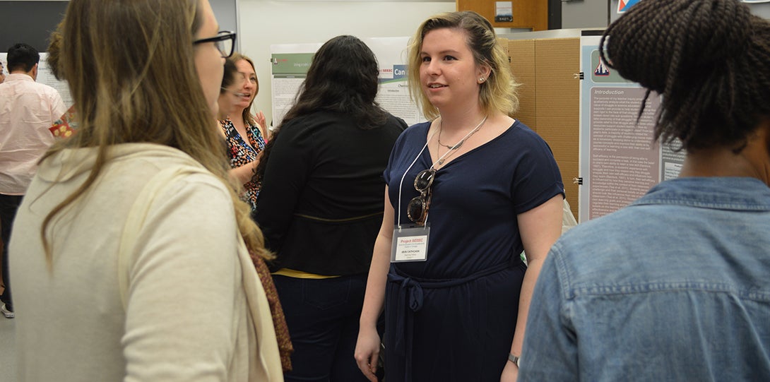 SEEEC Teaching Fellow Erin Cathcara talks with other fellows about her research project, with presentation posters behind her.