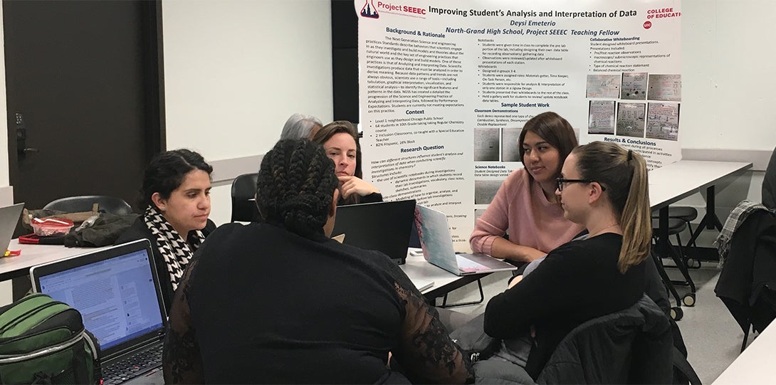SEEEC Teaching Fellow Deysi Emeterio sits at a table with other fellows, discussing her research.  Her poster presentation is behind her.