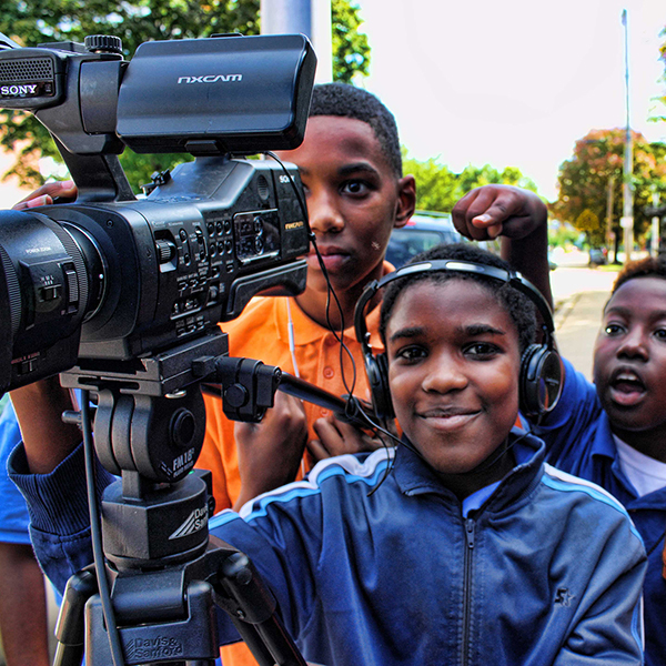Three young boys are manning a video camera, while one wears headphones, as they capture video in their community.